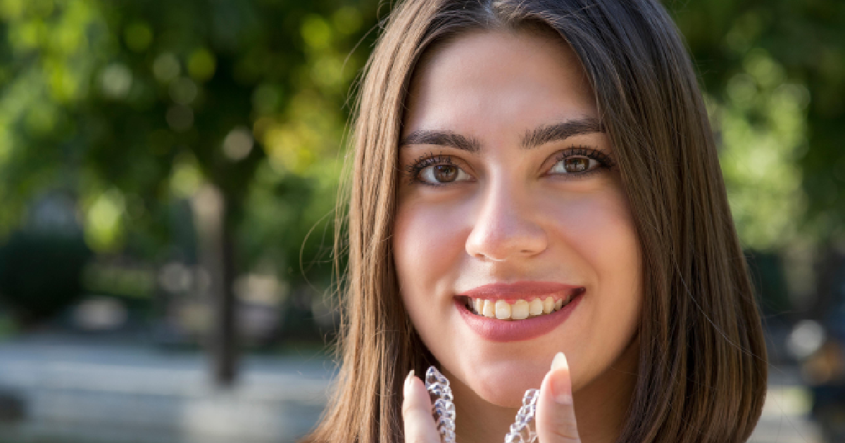 Smiling young woman holding a clear Invisalign aligner near her teeth, showcasing a natural smile outdoors with blurred greenery in the background, promoting Invisalign Treatment in Orland Park.